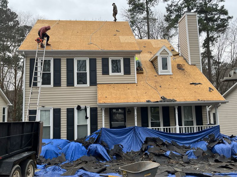 Roofers stripping old shingles from a house roof, exposing the plywood decking, with debris on blue tarps below