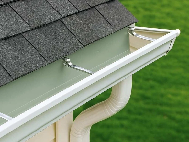 Gutter Installation — Newly installed white seamless gutter system with a downspout on a house with dark shingle roof, demons