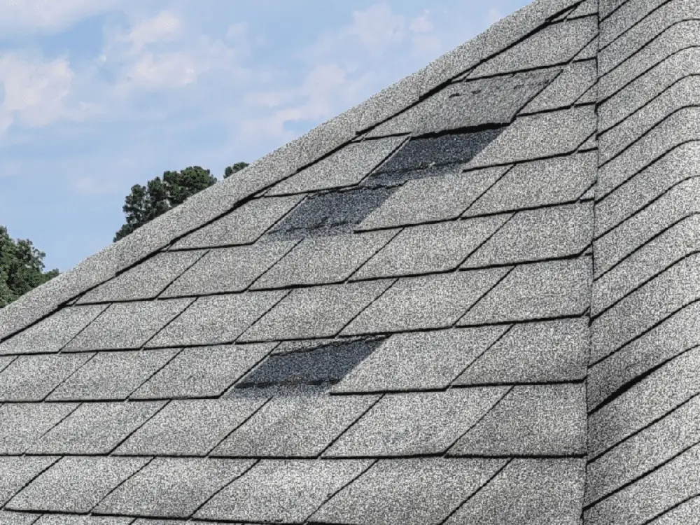 Side view of a gray shingle roof with multiple patches of missing shingles from wind damage, revealing darker underlayment