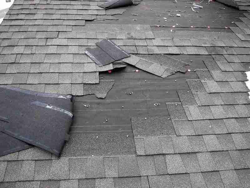 Extensive storm damage on a dark gray shingle roof with many shingles torn off and scattered, exposing the roof deck