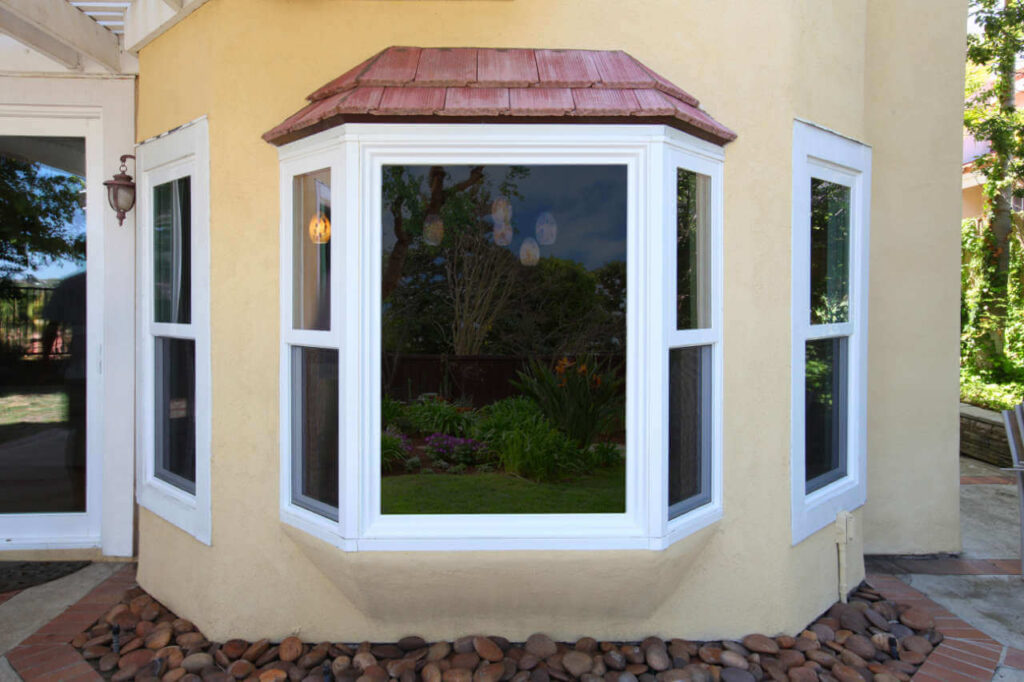 Exterior view of a bay window with white frames on a light yellow stucco house, surrounded by landscaping